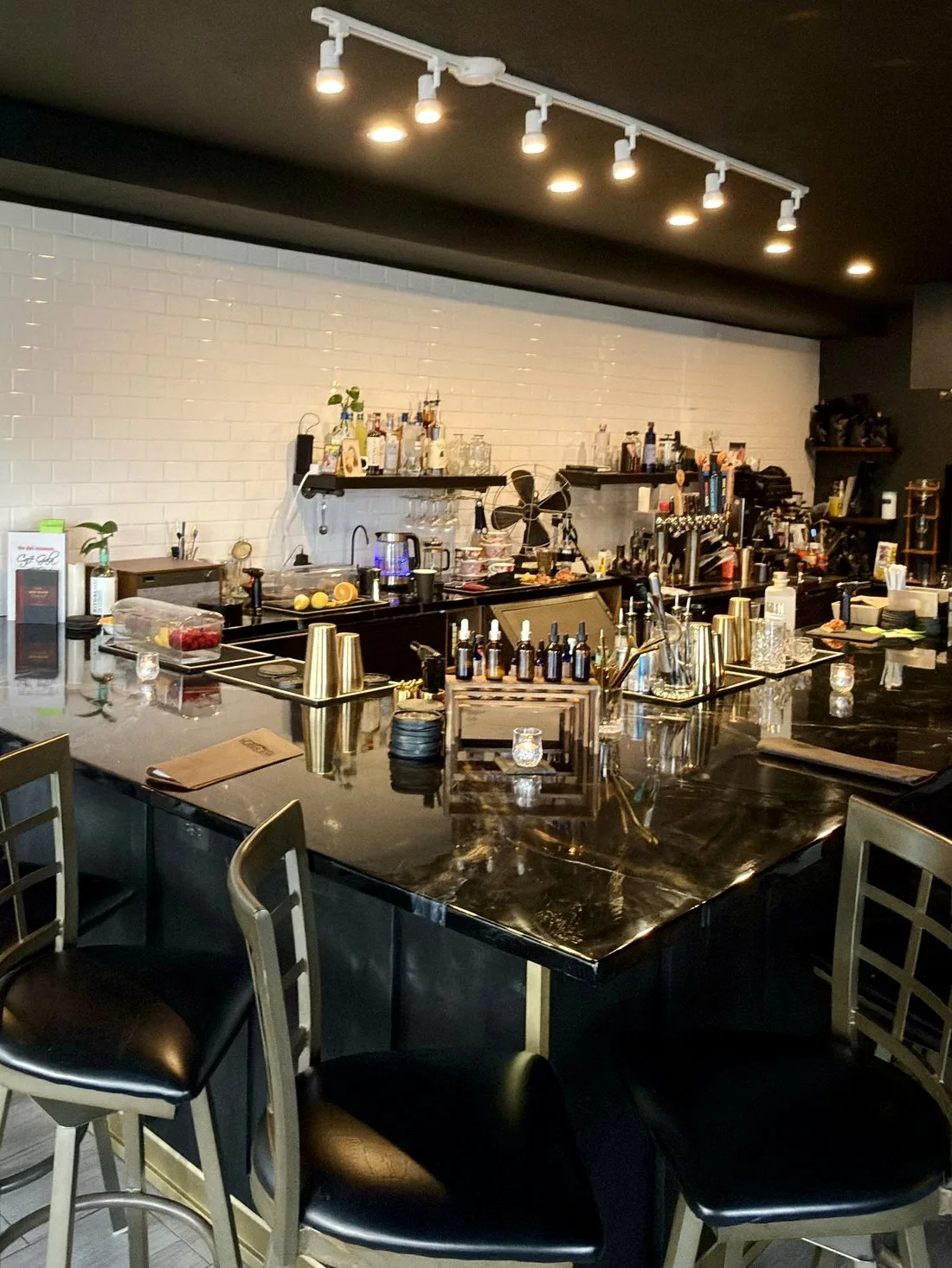 The black marble bar lined with brass barware and bitters, bottle shelves against white subway tile under warm track lighting